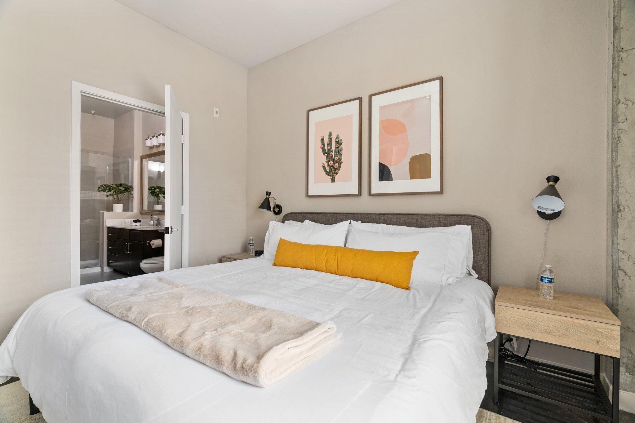 A neatly made bed with white and mustard pillows, a bedside table with a lamp, and two framed pictures on the wall.