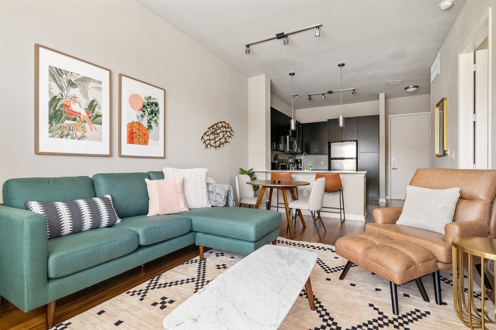 A modern living room with a teal sofa, a brown armchair, a marble coffee table, and a patterned rug. There are two framed artworks on the wall, a kitchen area in the background, and a dining table with chairs.
