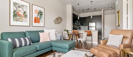 A modern living room with a teal sofa, a brown armchair, a marble coffee table, and a patterned rug. There are two framed artworks on the wall, a kitchen area in the background, and a dining table with chairs.