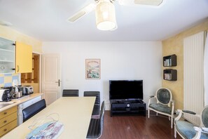 Ein Zimmer mit Esstisch, Stühlen, Fernseher, Bücherregal und Deckenventilator.