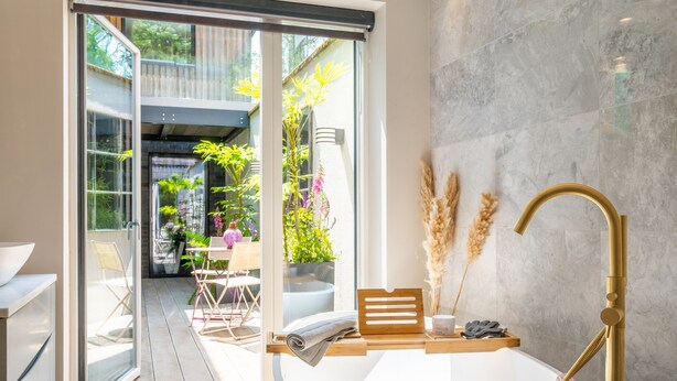 A modern bathroom with a freestanding bathtub, a glass door leading to an outdoor area with plants and seating, and a marble wall. - Gamekeeper's Lodge - sleeps 8 guests  in 4 bedrooms (Stratford-upon-Avon)