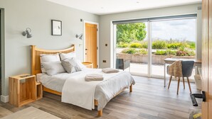 A modern bedroom with a wooden bed, a small wooden nightstand, a desk, and a chair. There is a large sliding glass door leading to a balcony with plants. - Gamekeeper's Lodge - sleeps 8 guests  in 4 bedrooms (Stratford-upon-Avon)