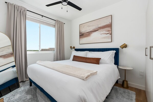 Quarto bem arrumado com cama grande, ventilador de teto, janela com cortinas, mesa de cabeceira e uma obra de arte na parede.