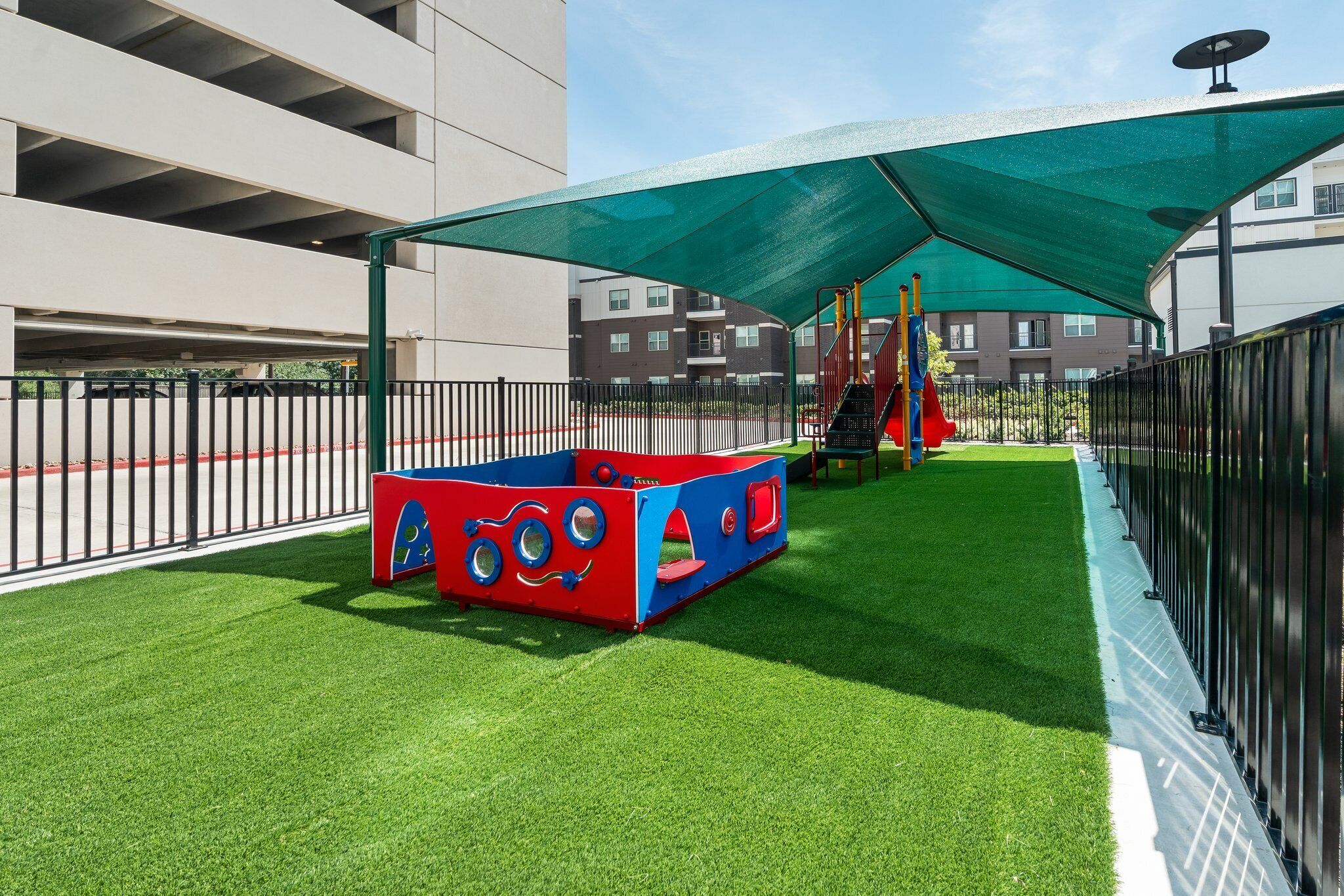 Ein Spielplatz mit einer roten und blauen Spielkonstruktion, grünem Kunstrasen und einem Sonnensegel, das Schatten spendet.