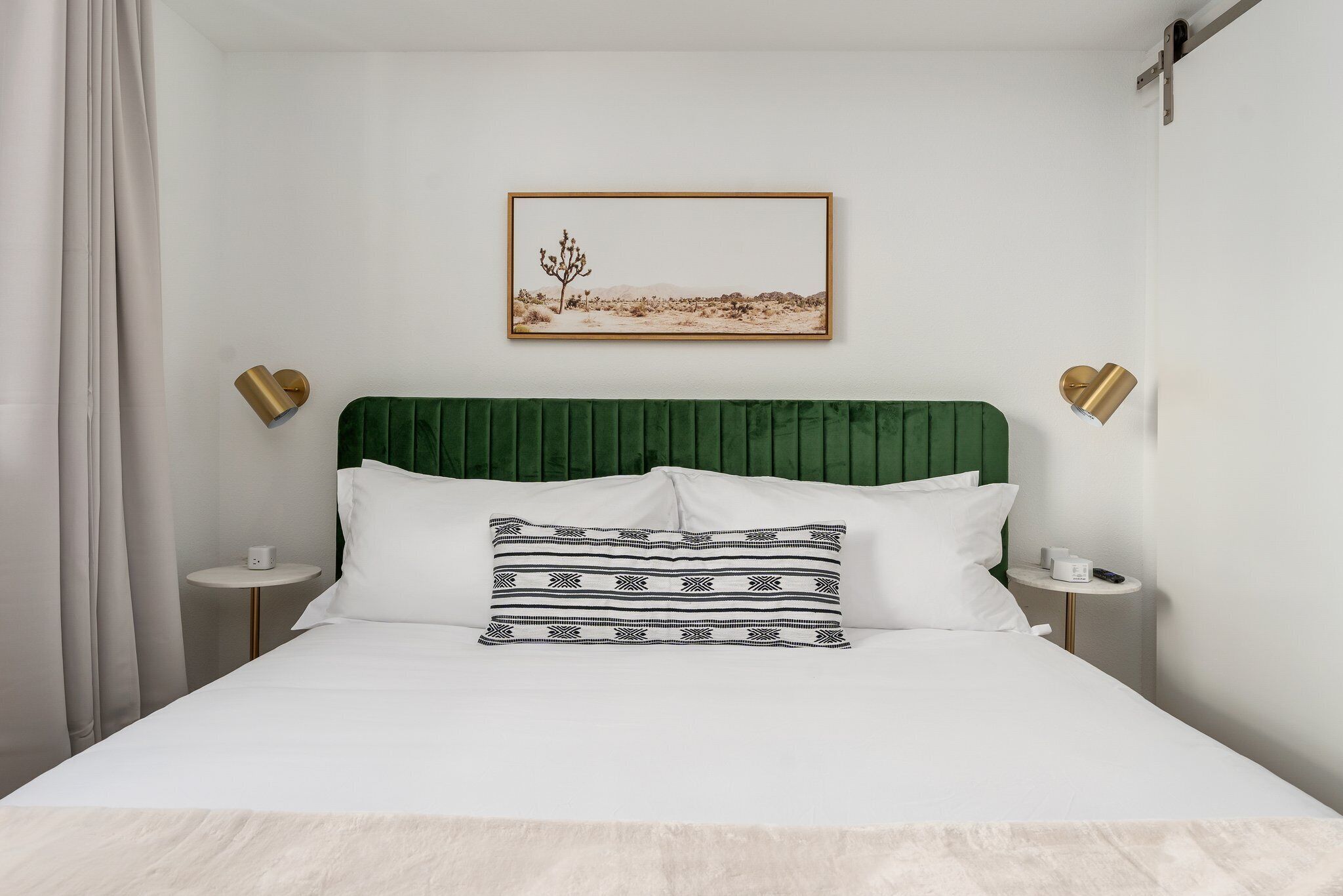 Cama bem arrumada com cabeceira verde, roupa de cama branca e almofada decorativa. Há um quadro na parede acima da cama e duas mesas de cabeceira com abajures.