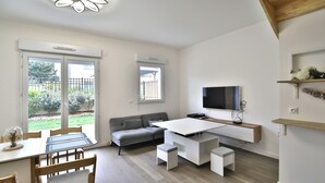 Un salon moderne comprenant un canapé, une table à manger avec des chaises, un téléviseur fixé au mur et une fenêtre donnant sur l’extérieur.