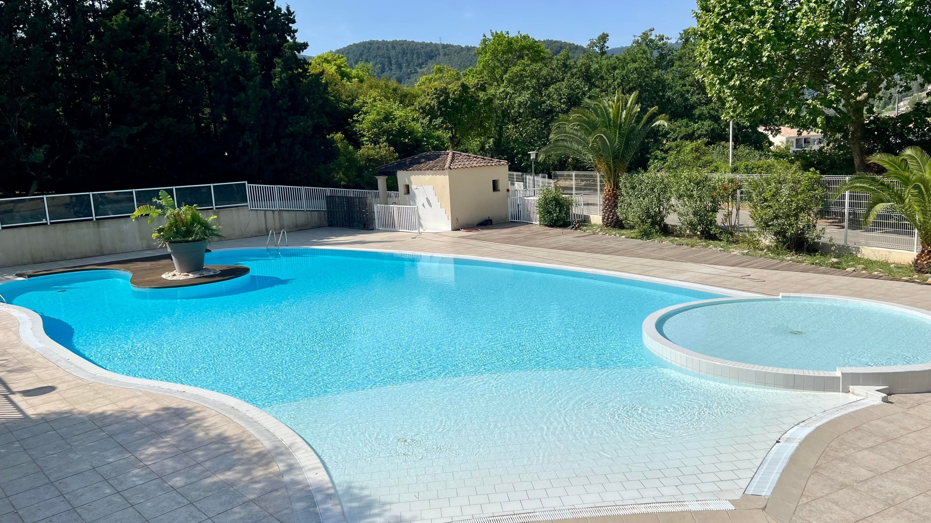 A swimming pool with a tiled deck, surrounded by greenery and a small building.