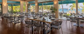 A spacious outdoor dining area with multiple tables and chairs, large windows offering a view of the ocean and palm trees. - Bayside Retreat - Walk to Cowpet Beach, Pool w/ Grotto, Oceanview! (St Thomas)