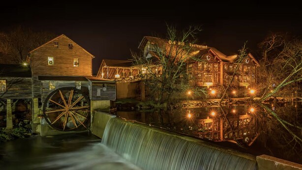 Un bâtiment historique doté d’une grande roue à eau, une structure en bois sur laquelle on peut lire l’inscription « Restaurant de l’Ancien Moulin », et dont l’image se reflète dans l’eau.