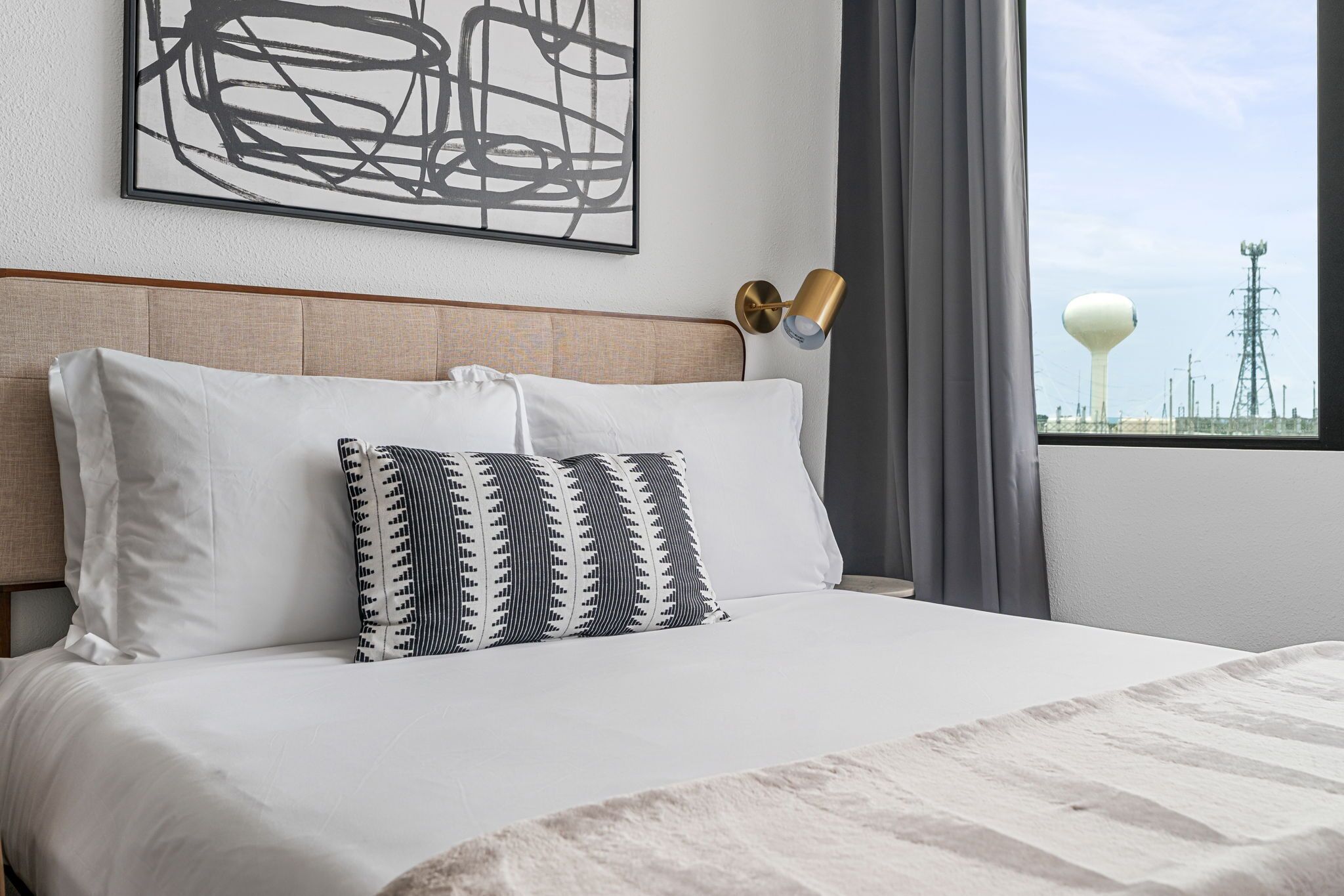 Een opgemaakt bed met witte lakens en gestreepte kussens, een modern abstract schilderij aan de muur, en zicht op een watertoren en hoogspanningslijnen door een raam.