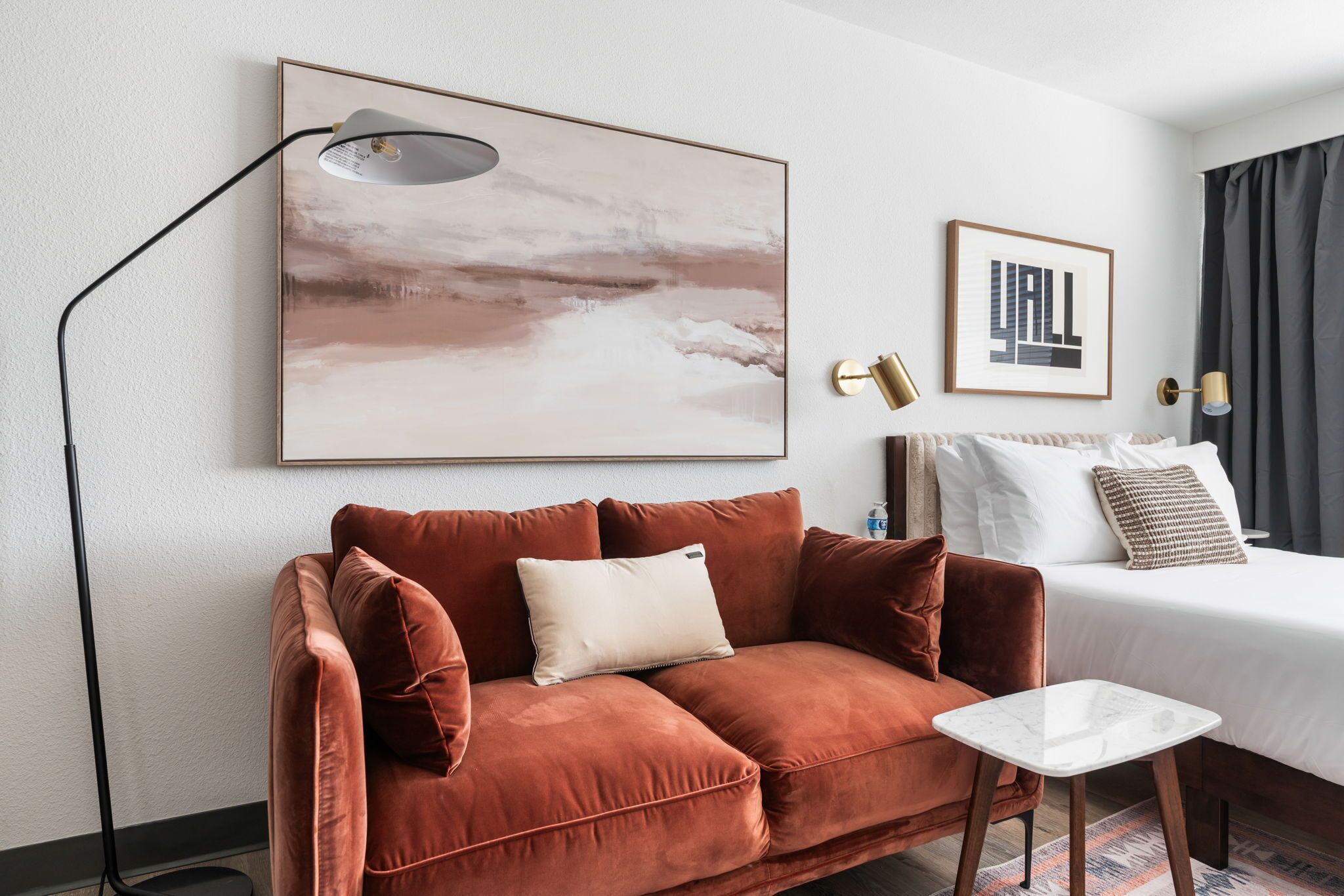 A hotel room with a brown sofa, a small white table, a bed with white bedding, a framed artwork, and a floor lamp.