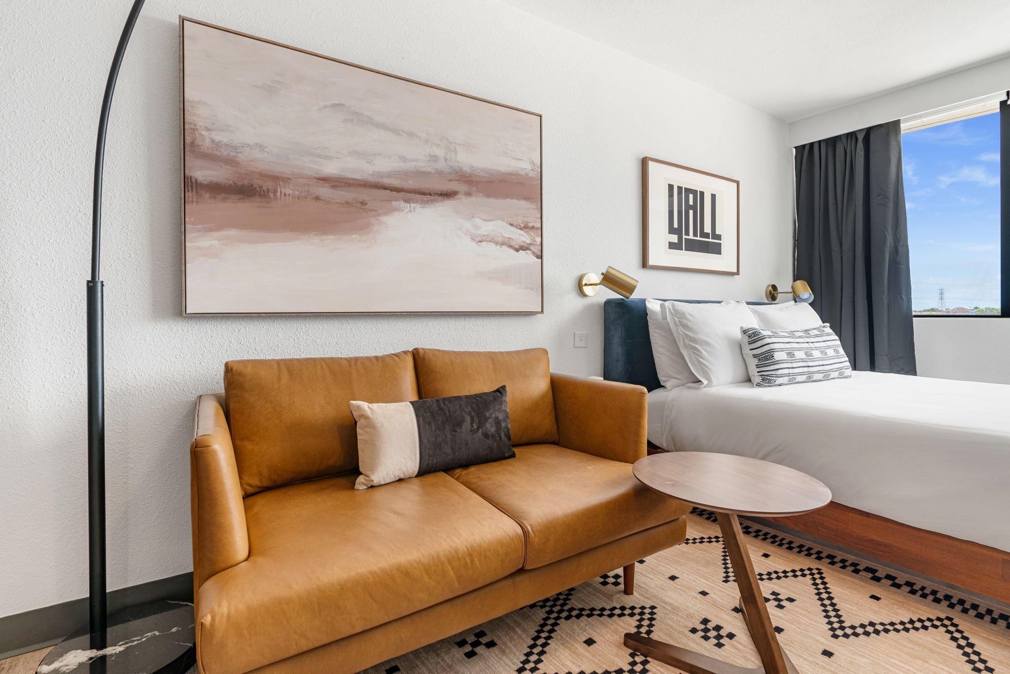 A modern hotel room with a leather sofa, a small round table, a bed with a white comforter, a large abstract painting, and a framed picture on the wall.