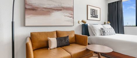 A modern hotel room with a leather sofa, a small round table, a bed with a white comforter, a large abstract painting, and a framed picture on the wall.