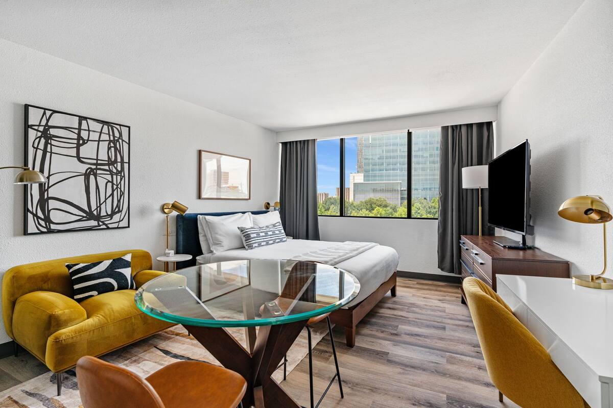 A modern hotel room with a bed, a glass table, a sofa, a TV, and a city view.