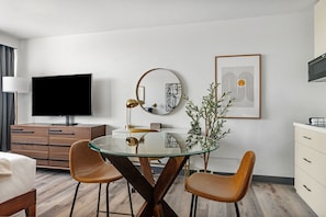 A modern hotel room with a glass dining table, two brown chairs, a flat-screen TV, a round mirror, and a framed abstract art piece on the wall. - Lodgeur Upper Kirby (Houston)