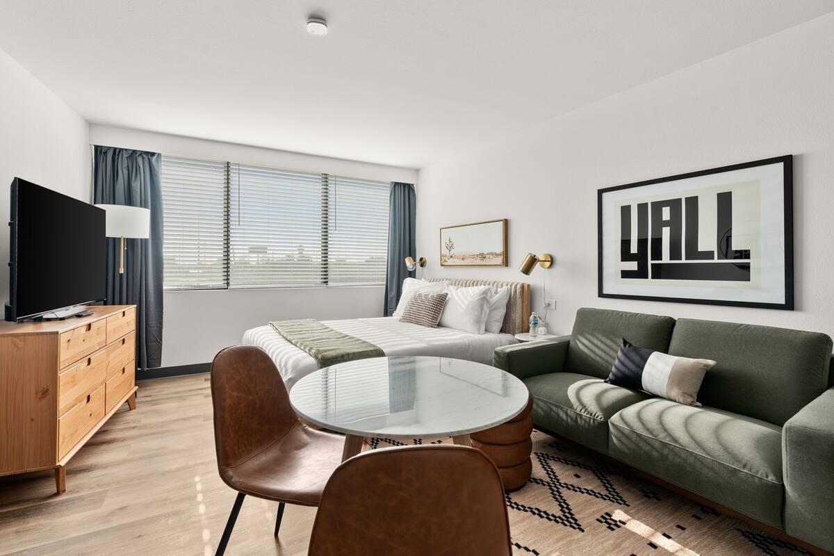 A modern hotel room with a bed, a green sofa, a round glass table, and a TV.