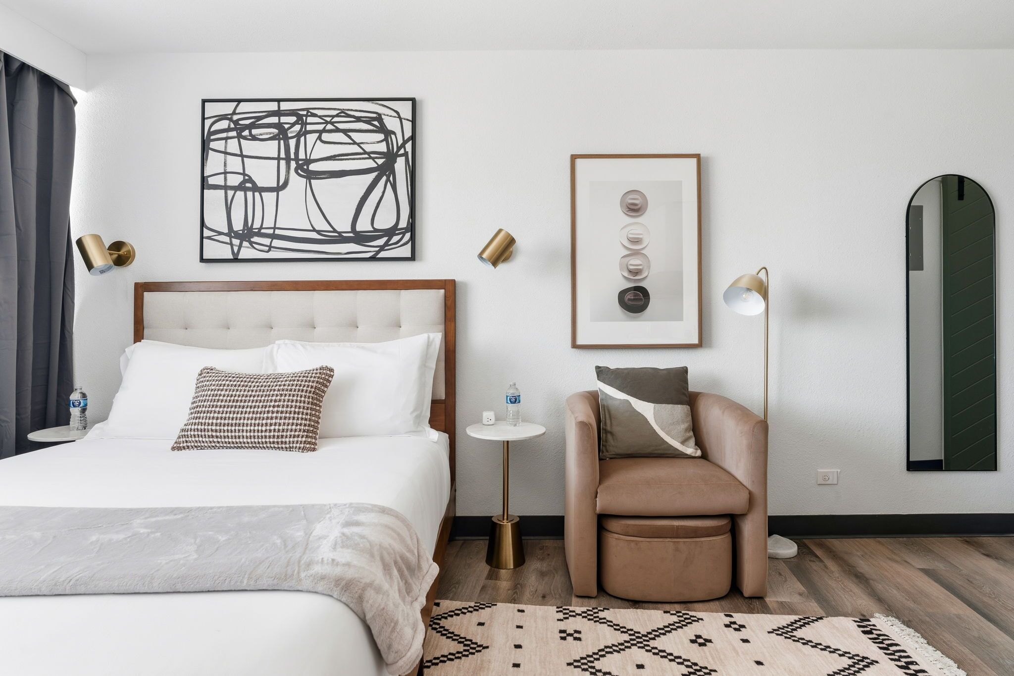 A hotel room with a bed, a chair, a small table with a water bottle, a framed picture, and a mirror.
