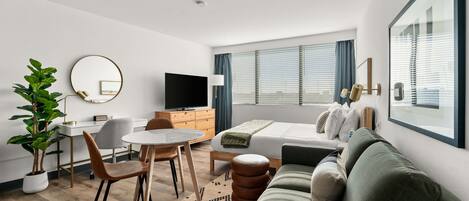 A modern hotel room with a bed, a sofa, a dining table with chairs, a TV, and a large window with blinds.