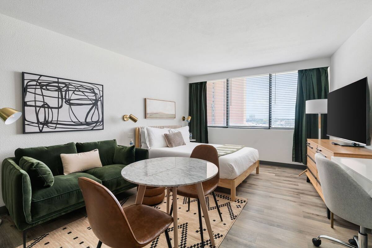 A modern hotel room with a green sofa, a round table, a bed, a TV, and a window with curtains.