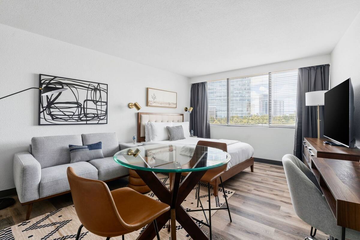 A modern hotel room with a glass dining table, a sofa, a bed, a TV, and a large window with a city view.