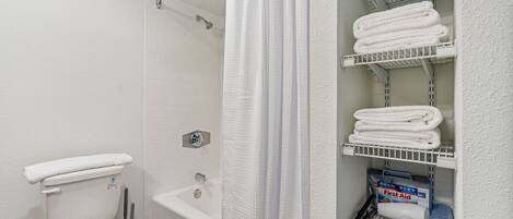 A bathroom with a toilet, shower, and shelves with towels and toiletries.