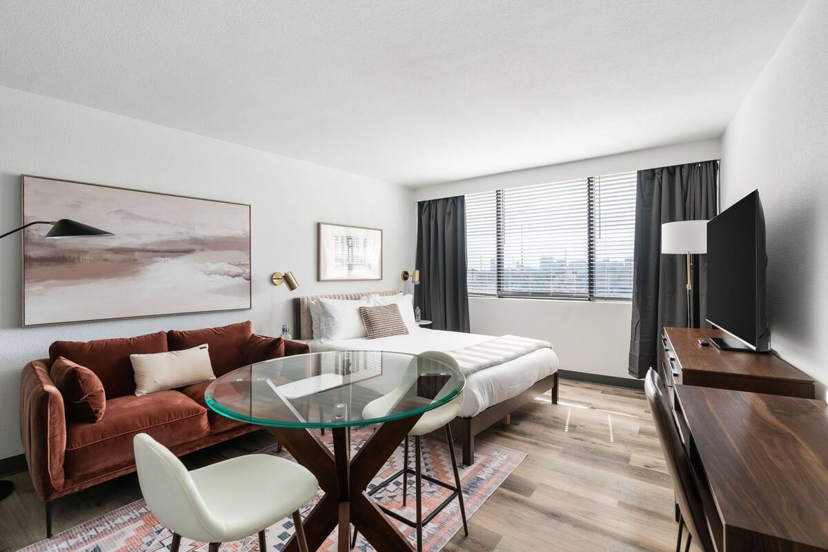 A modern hotel room with a glass dining table, a sofa, a bed, and a TV.