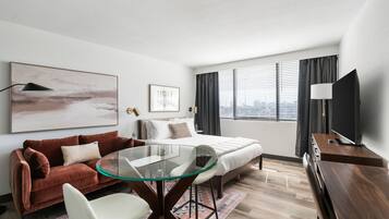 Ett modernt hotellrum med en säng, ett glasbord, en soffa, en TV och utsikt över staden.