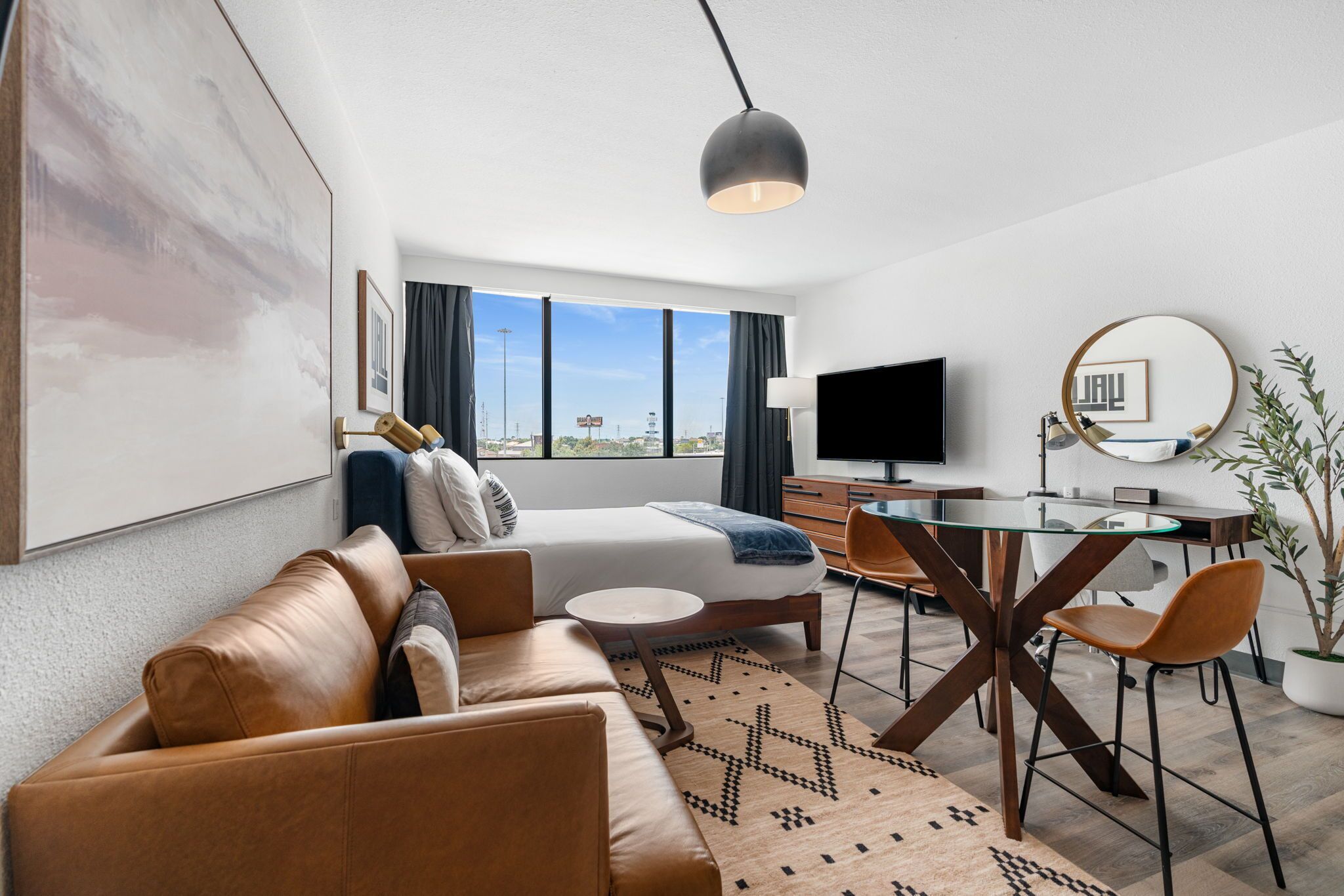 A modern hotel room with a large bed, a sofa, a dining table with chairs, a TV, and a large window with a city view.