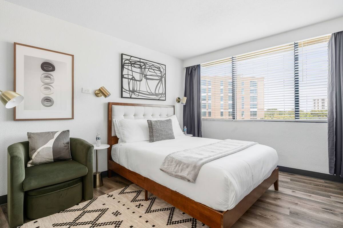 A modern hotel room with a large bed, a green armchair, a small side table, and a view of the city through large windows.