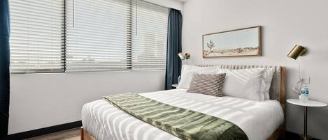 Cama bem arrumada com manta verde, quadro na parede e mesa de cabeceira com luminária e garrafa de água.