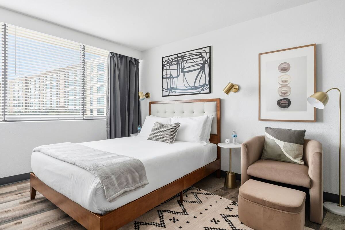 A modern hotel room with a large bed, a side table, a chair, and a window with blinds.