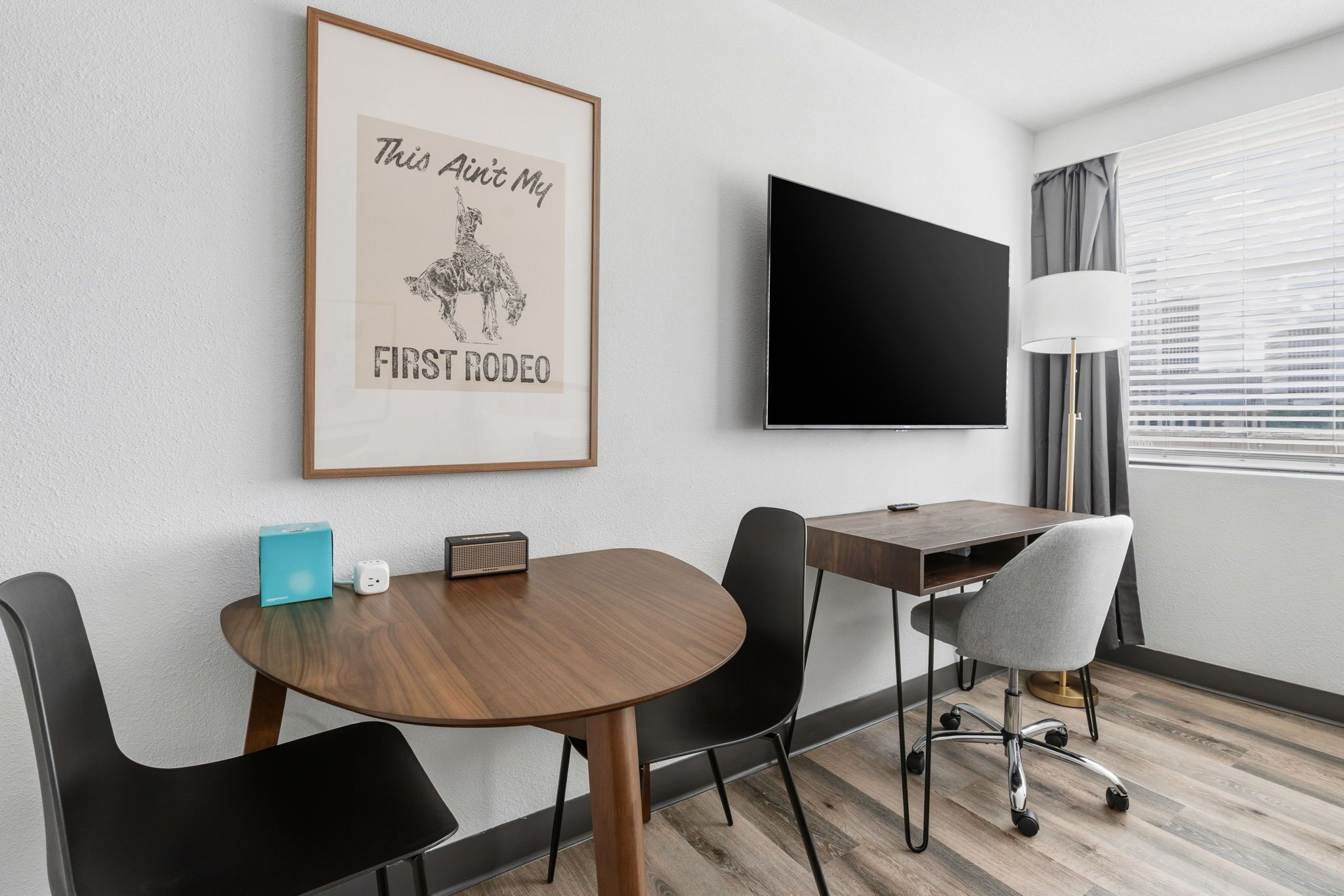 Ein Hotelzimmer mit einem Holztisch, schwarzen Stühlen, einem Schreibtisch, einem Flachbildfernseher, einem gerahmten Poster, einer Lampe und einem Fenster mit Jalousien.