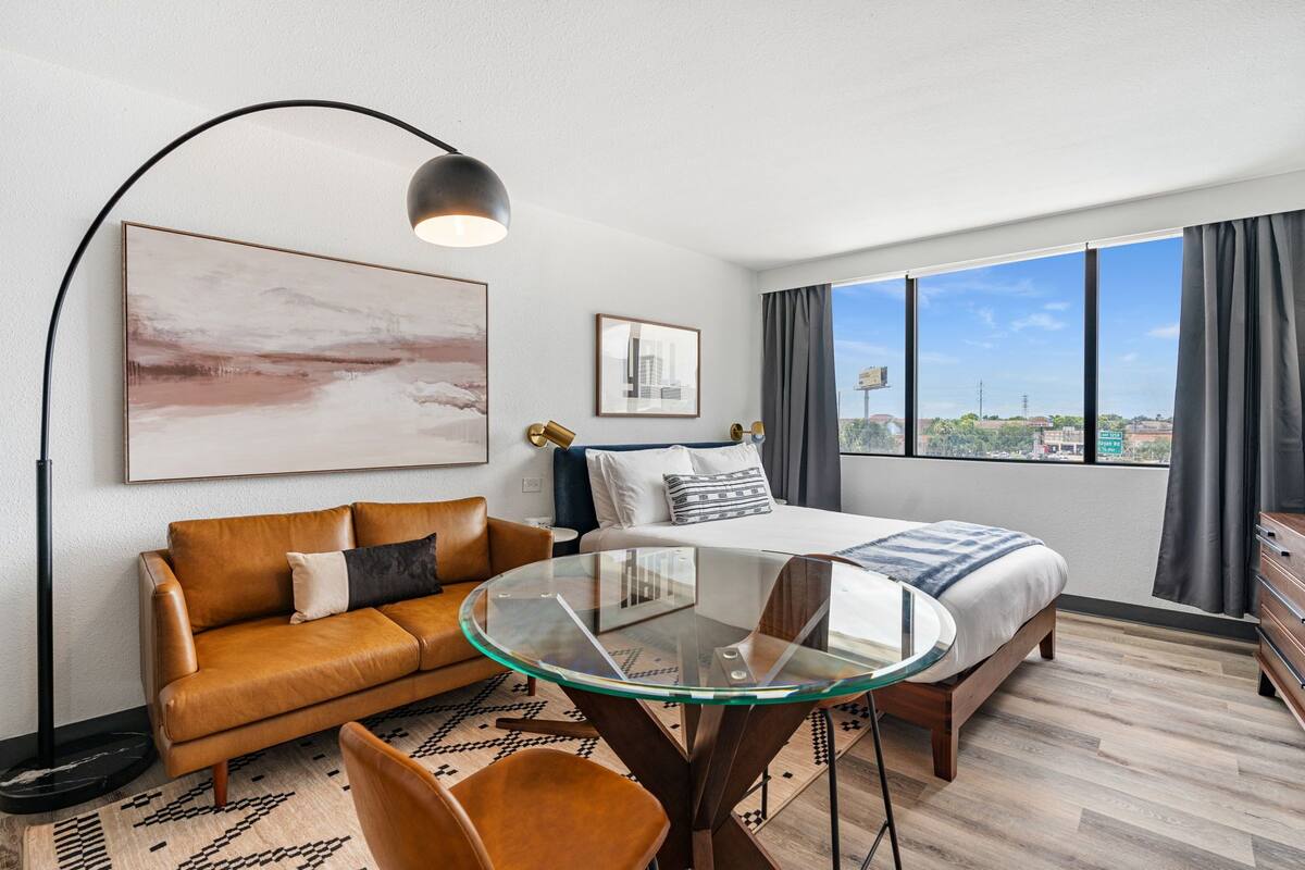 A modern hotel room with a bed, a glass table, a sofa, and a large window offering a city view.