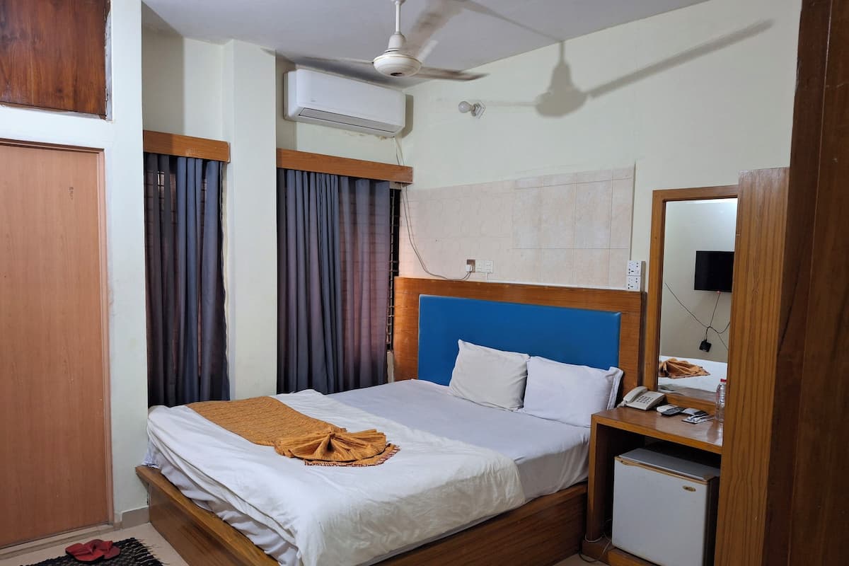 Ein Hotelzimmer mit einem hölzernen Bett, einem kleinen Nachttisch mit Telefon und Flasche, einem Deckenventilator und einer Klimaanlage.