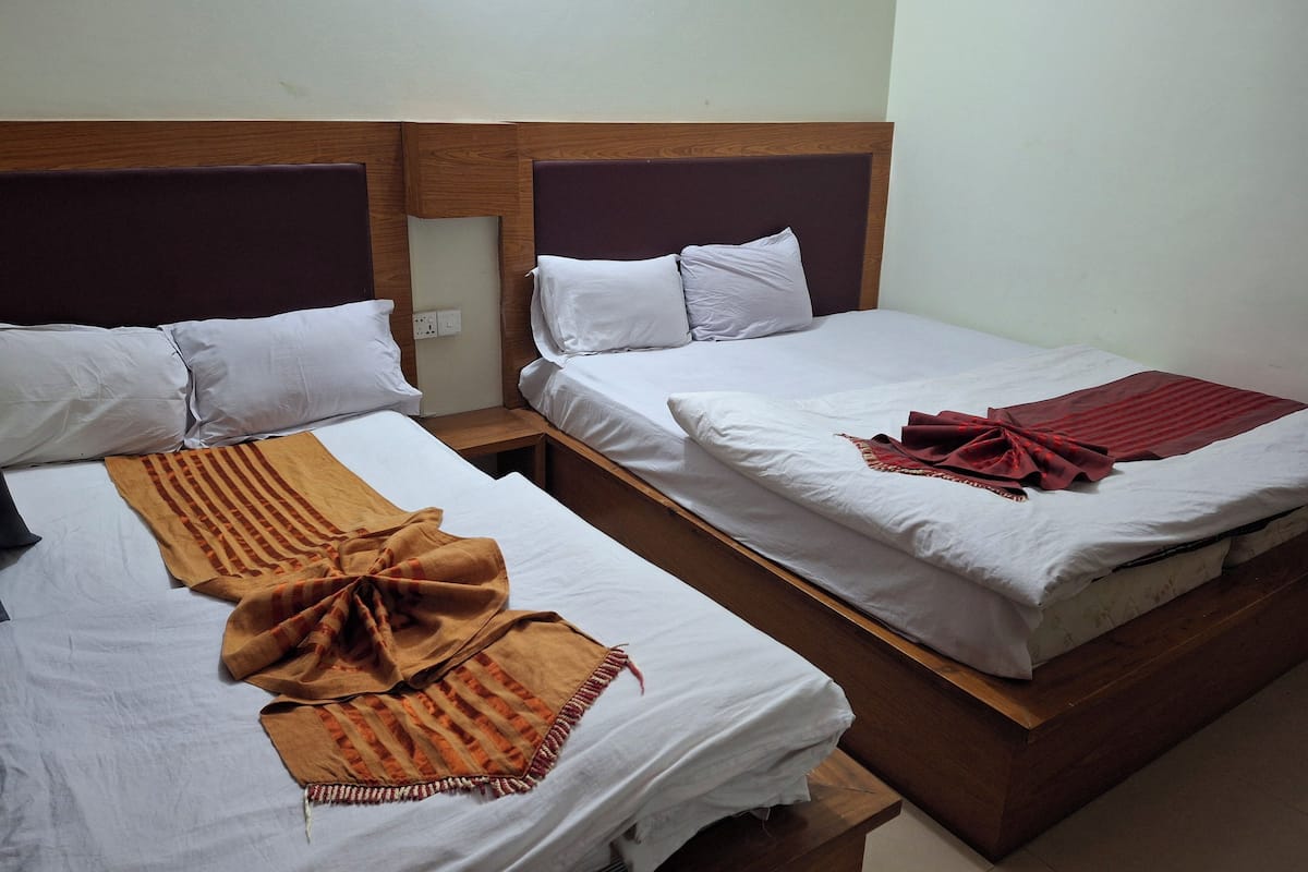 Ein Hotelzimmer mit zwei Einzelbetten, jeweils mit weißer Bettwäsche und einem Holz-Kopfteil. Auf einem Bett liegt eine braun gestreifte Decke, auf dem anderen eine rote Decke.