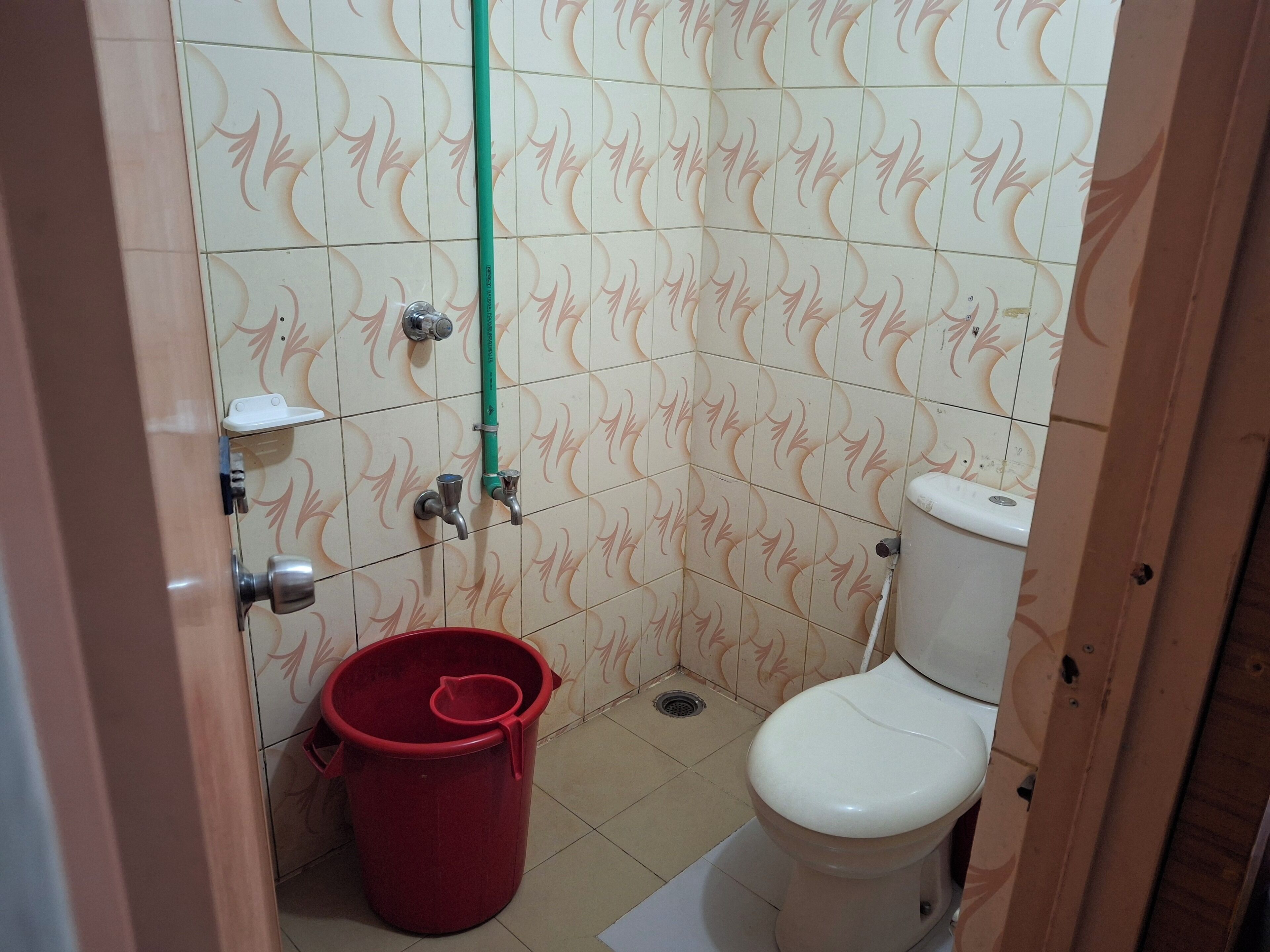 A small bathroom with a toilet, a red bucket, and a green pipe.