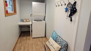 Een compacte kamer met een kitchenette, een bank met kussen, een houten lepel aan de muur en een brandblusser in het raam.