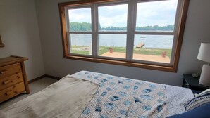 Een slaapkamer met een groot raam met uitzicht op een meer, een bed met een blauw-witte quilt, een houten dressoir en een nachtkastje met een lamp.