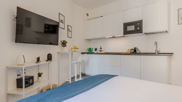 Une chambre moderne et compacte, équipée d’un lit, d’une petite kitchenette, d’un téléviseur à écran plat et d’un bureau avec une chaise.