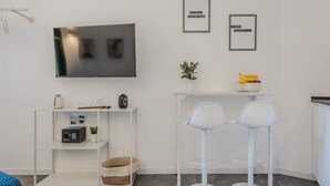 Un espace de vie moderne et minimaliste, doté d’un téléviseur à écran plat, de deux tabourets de bar blancs, d’une petite table blanche sur laquelle se trouve un saladier rempli de fruits et une plante, ainsi que d’une étagère blanche contenant divers objets.
