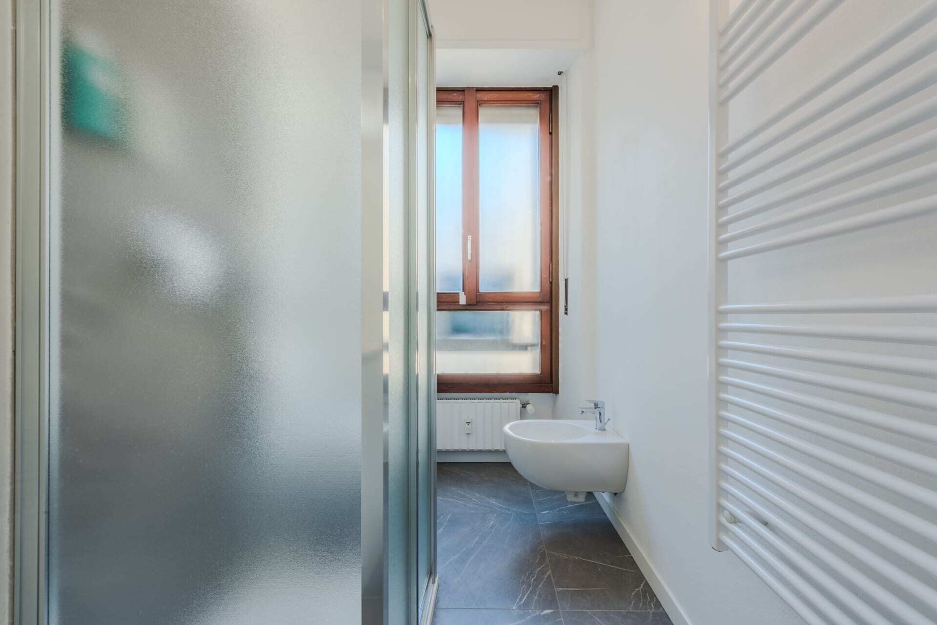 A narrow hallway leading to a bathroom with a window, a radiator, and a bidet.