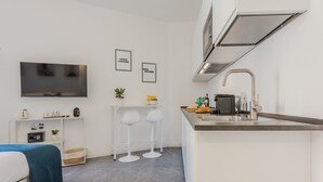 Une cuisine moderne équipée d’une cuisinière intégrée, d’un évier et d’un téléviseur à écran plat fixé au mur. On y trouve deux tabourets de bar blancs et une petite table blanche agrémentée d’une plante et d’un saladier de fruits.