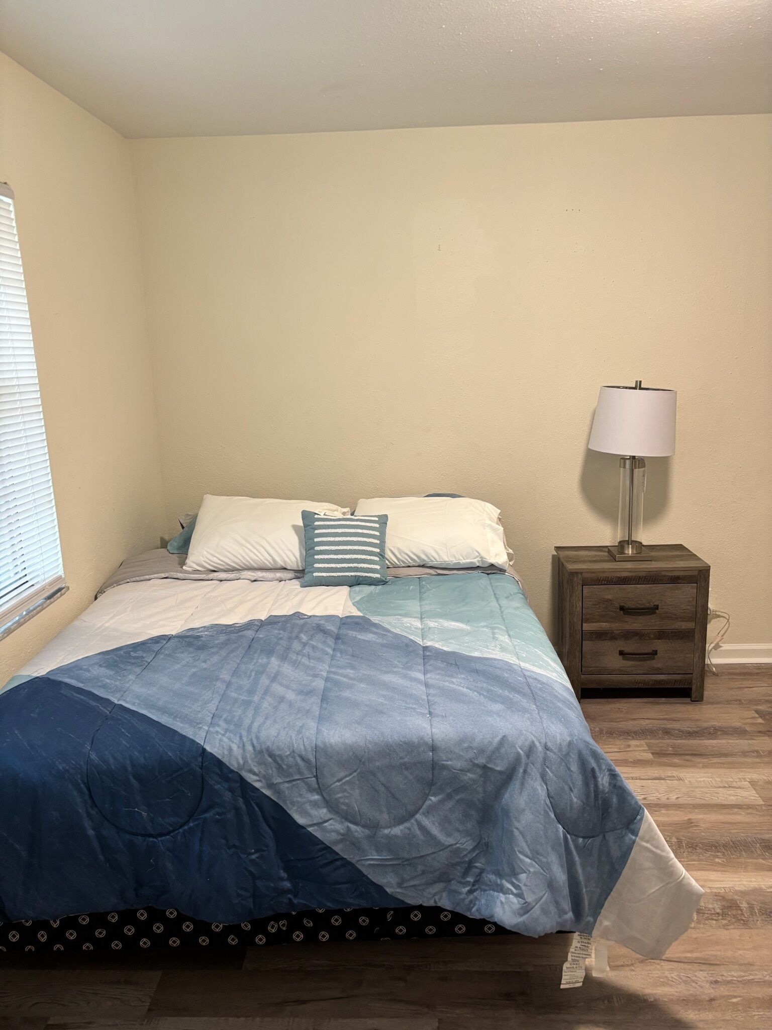 A bedroom with a bed, two pillows, a nightstand with a lamp, and a window with blinds.