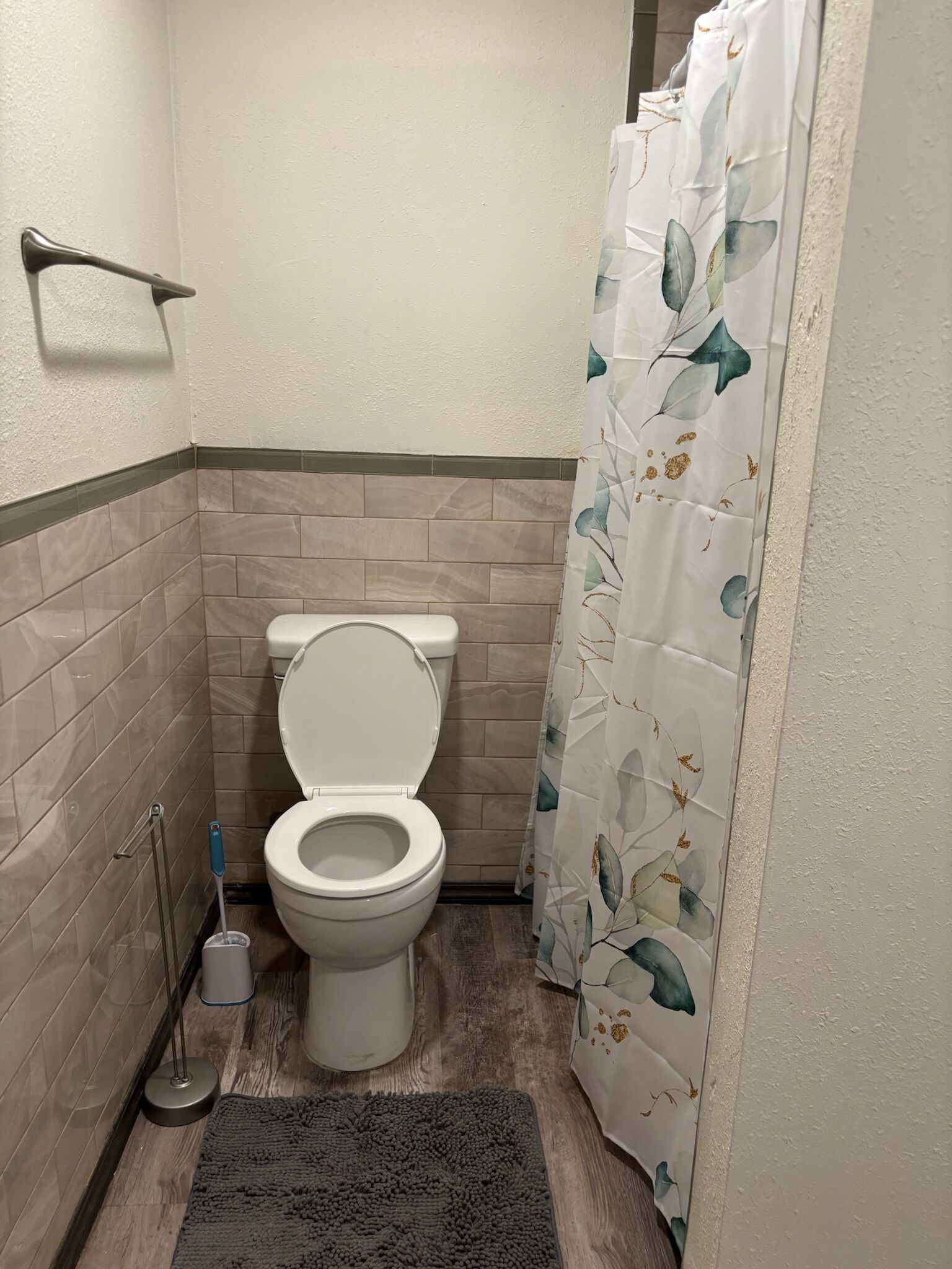 Een badkamer met toilet, douchegordijn met bladmotief, vloermat en een handgreep.