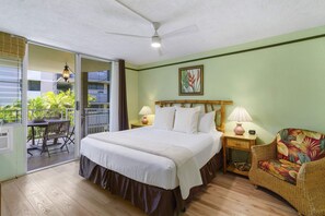A hotel room with a bed, a nightstand, a chair, a balcony with a table and chairs, and a ceiling fan.