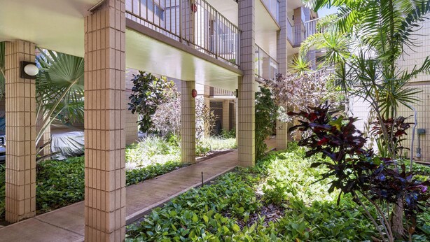 A covered outdoor area with a pathway, surrounded by greenery and a building with balconies.