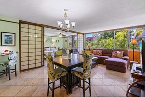 Un espace repas comprenant une table et des chaises, un canapé et une vue sur un paysage tropical grâce à de grandes fenêtres.