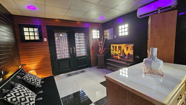 A modern living room with a black and white checkered floor, a black sofa with patterned cushions, a wooden door with glass panels, and a television displaying a room scene. - The Studio of Praise - by Zenbreak (Claxton Bay)