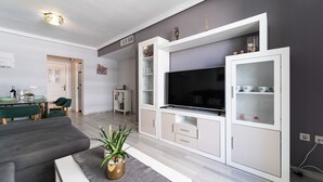 A modern living room with a flat-screen TV, built-in shelving, and a dining area with green chairs and a table. - MalagaSuite Sky & Sea Torremolinos (Torremolinos)