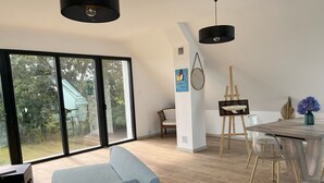 A modern living room with a large sliding glass door leading to a garden, a dining area with a wooden table and chairs, and a blue sofa.